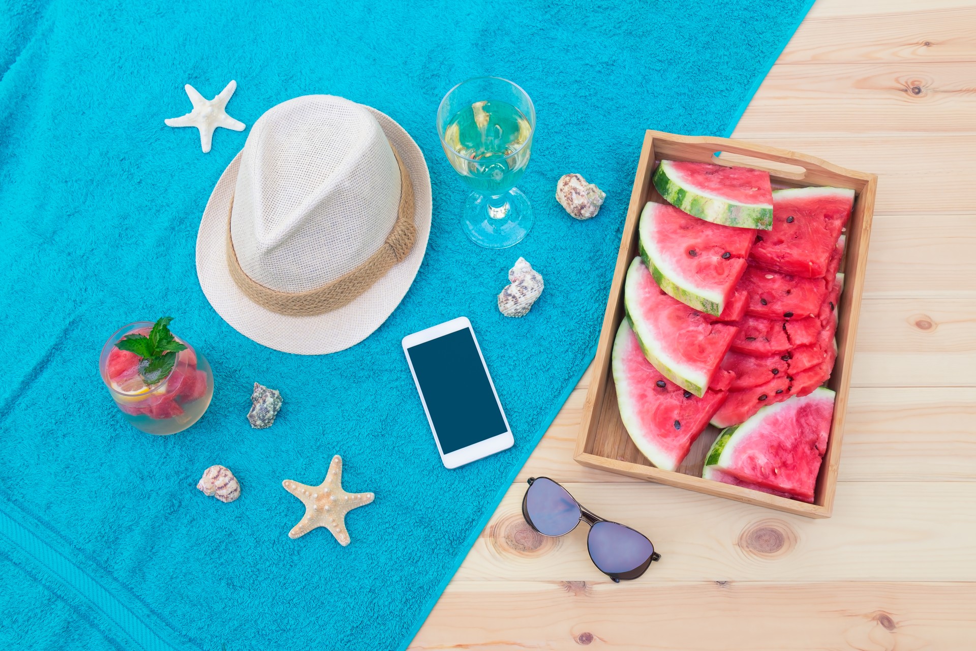 Hat, smartphone, starfishes, seashells, white wine, watermelon, watermelon lemonade and sunglasses on towel on wooden surface. Top view.